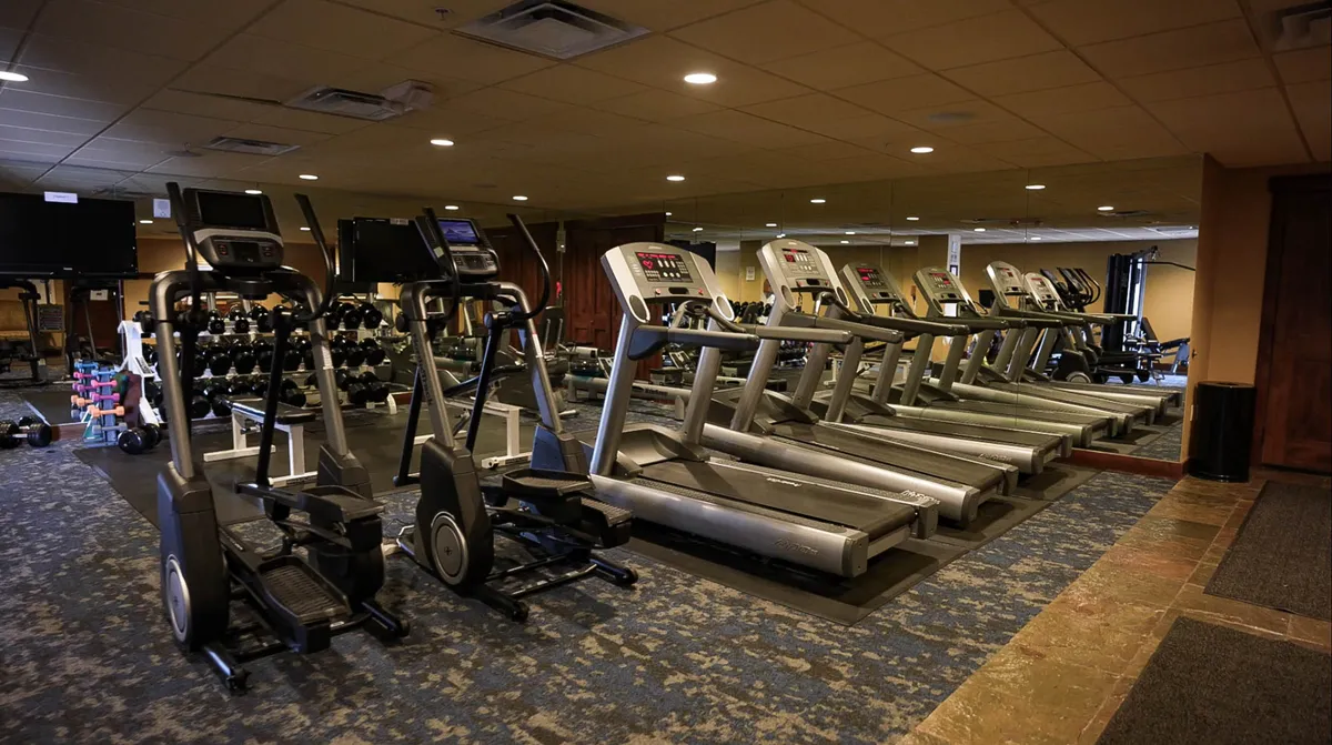 An interior view of the fitness center, showing treadmills, ellipticals, and a range of free weights. The setup is modern and spacious, emphasizing personal wellness and the social aspect of working out.