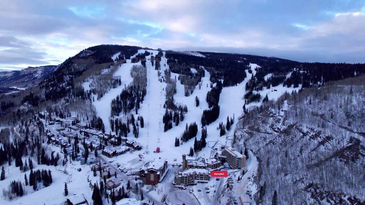 A sweeping view of ski slopes bordered by pine trees, with residential buildings nestled at the base. Chair lifts ascend the mountain, inviting visitors to explore the snowy terrain. The landscape captures the thrill of winter sports with a serene natural backdrop.