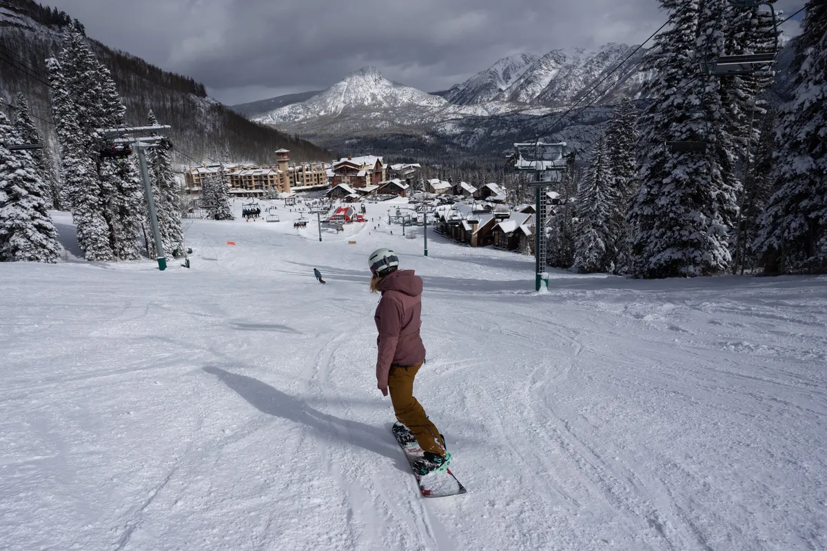 A snowboarder carves down a powdery slope under a sunrise sky. The scene includes a ski lift, a snow-covered mountain range, and a cozy village below, blending excitement and relaxation within a single frame.