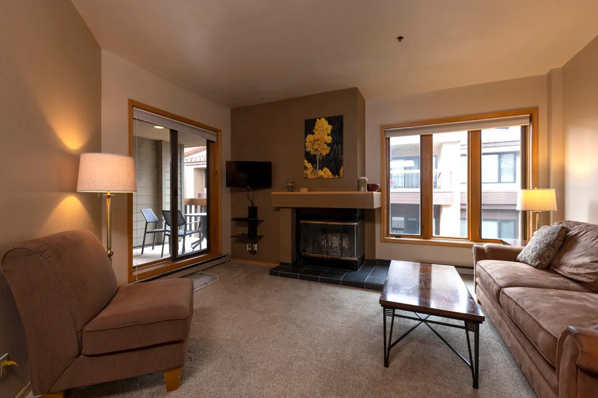 The image showcases a cozy living area within the condo, featuring a glowing wood-burning fireplace as the focal point. Large windows flood the space with natural light, while comfortable seating—including a plush sofa and inviting armchair—creates the perfect spot for relaxation. Artwork on the walls adds a touch of warmth and personality, and a coffee table completes the inviting setup, ideal for unwinding after a day on the mountain.
