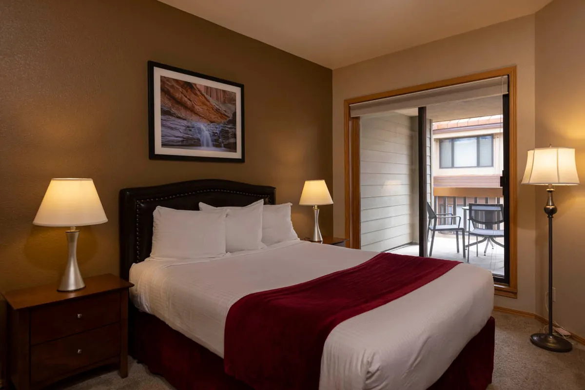A comfortable bedroom interior featuring a bed with white linens and a red accent blanket, flanked by nightstands with lamps and framed artwork. A balcony door lets in soft natural light, creating a tranquil retreat-like atmosphere.