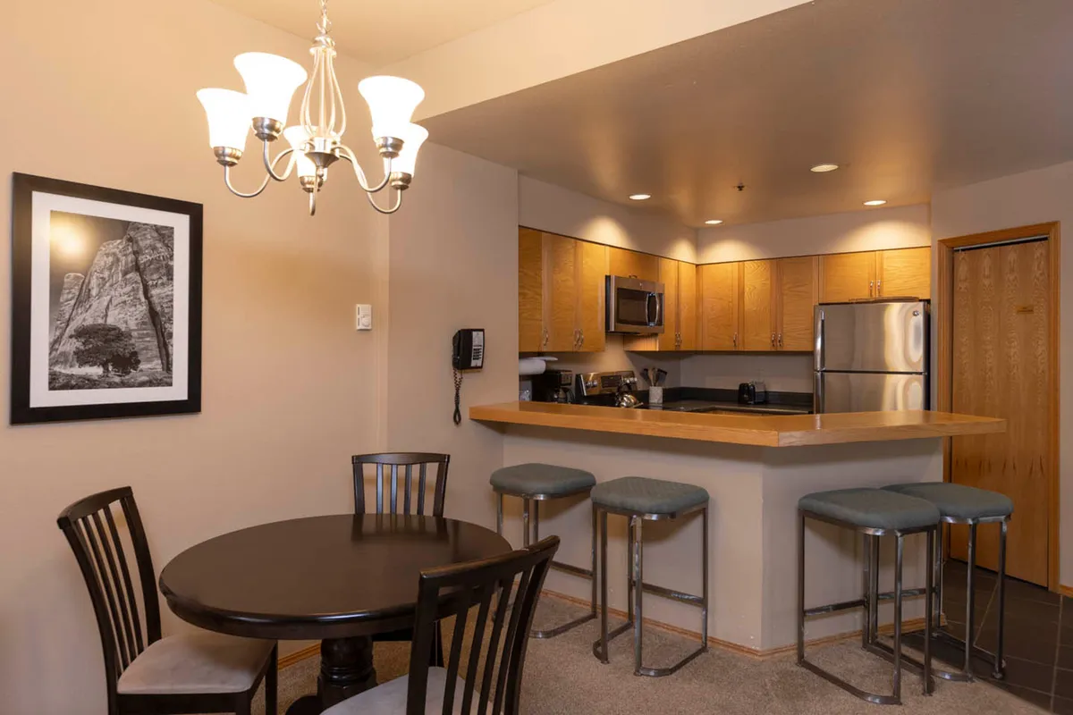 A welcoming interior scene centered around a round dining table with a chandelier overhead, a bar counter, and visible kitchen appliances. The space exudes warmth and is arranged for entertaining and relaxing, with touches like framed photographs adding a personal feel.